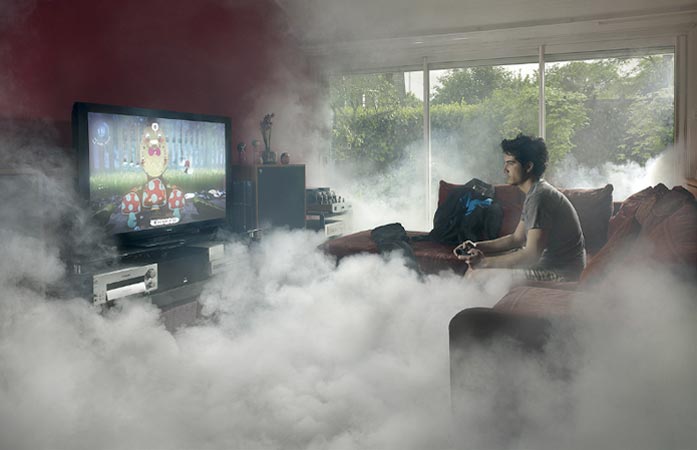 Teenage boy playing video game on TV with controller, sitting on couch in a room filled with artificial fog; entertainment scene.