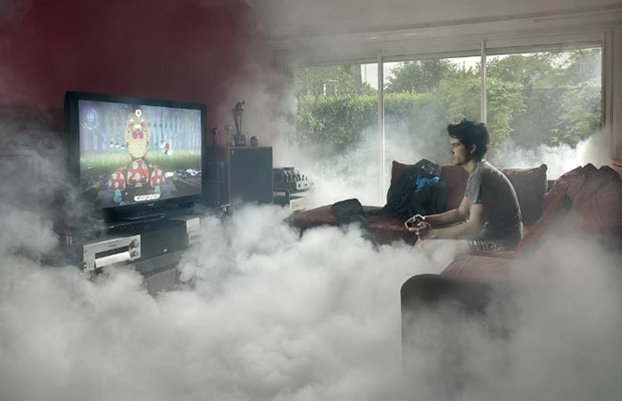 Teenage boy playing video game on TV with controller, sitting on couch in a room filled with artificial fog; entertainment scene.