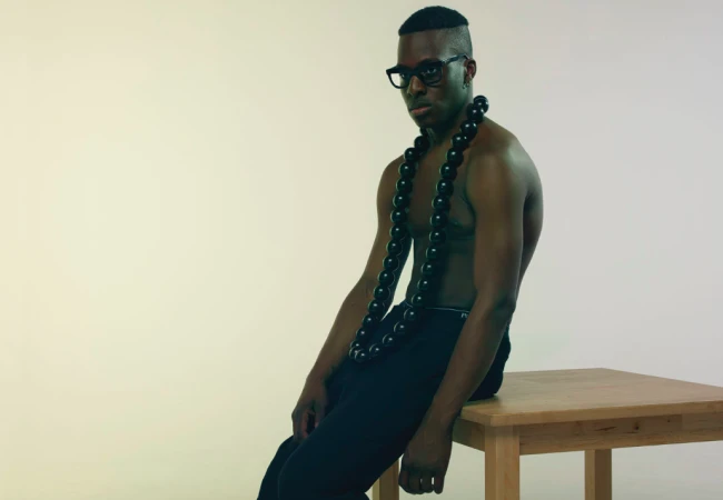 Shirtless black man wearing thick black framed glasses and a large beaded necklace, seated on a wooden table against a white background