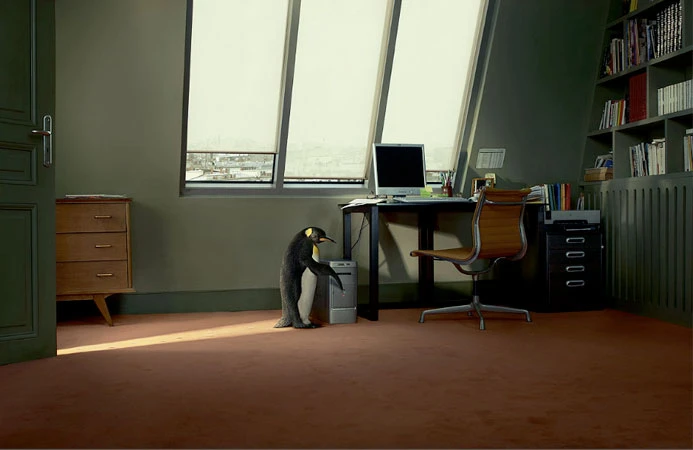 A lone penguin stands in an office next to a computer tower under a window, creating a surreal and humorous office scene.
