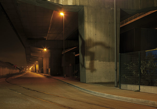 A street under an overpass at night illuminated by streetlights creates an eerie and industrial landscape with concrete and shadows.