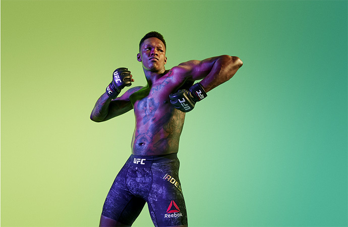 Shirtless fighter Izzy Adesanya posing in a studio with boxing gloves, tattoos, and Reebok trunks against a green gradient backdrop.