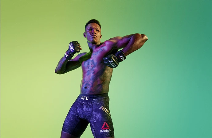 Shirtless fighter Izzy Adesanya posing in a studio with boxing gloves, tattoos, and Reebok trunks against a green gradient backdrop.