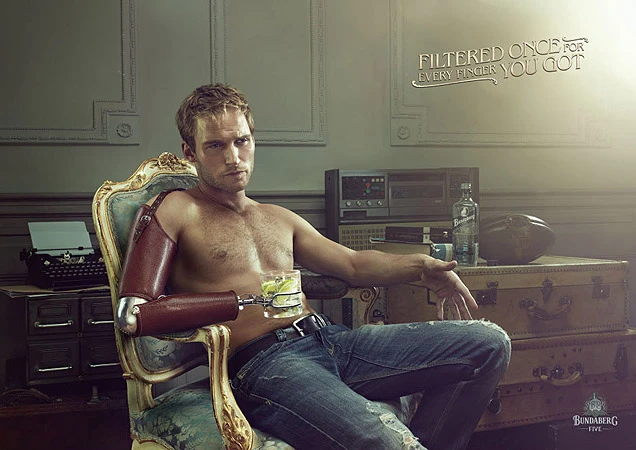 Shirtless man with prosthetic arm sits with a cocktail near a vintage typewriter and stereo in dim, atmospheric light.