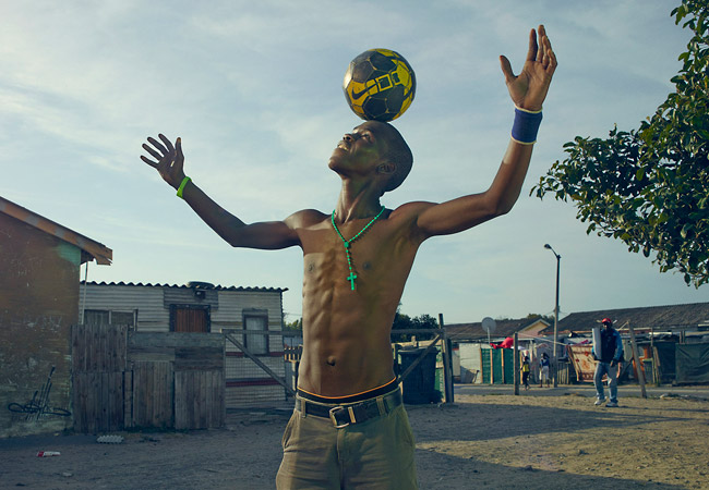 Athletic young man balances a soccer ball on his head in an outdoor setting, wearing casual clothing with simple buildings visible.