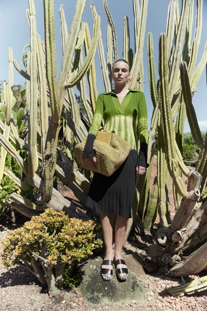A woman poses outdoors wearing a green tie-dye shirt, black skirt, and carrying a woven tote bag, against a backdrop of tall cacti.