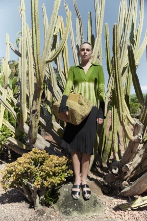 A woman poses outdoors wearing a green tie-dye shirt, black skirt, and carrying a woven tote bag, against a backdrop of tall cacti.