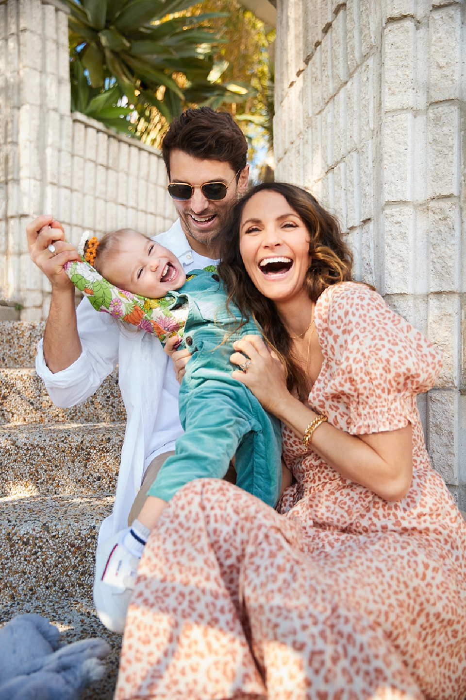 A happy family of three poses outside on a sunny day. A man holds up a laughing baby, while a smiling woman sits next to them.