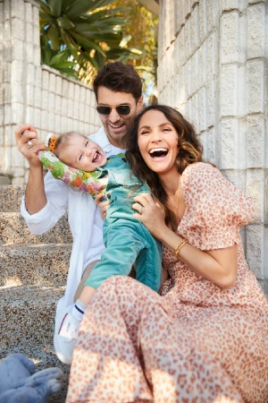 A happy family of three poses outside on a sunny day. A man holds up a laughing baby, while a smiling woman sits next to them.