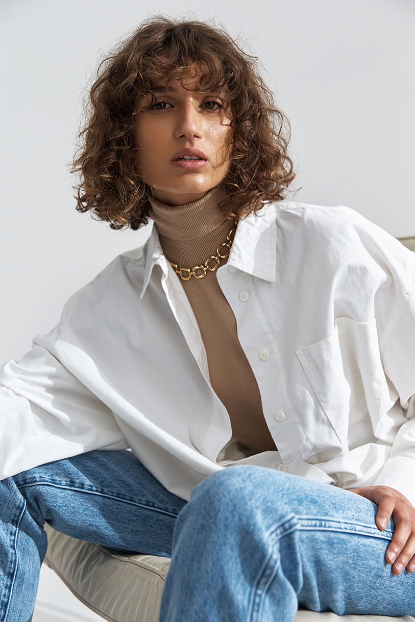 Attractive brunette woman in a white button-down shirt and blue jeans poses in a bright studio with a gold necklace.