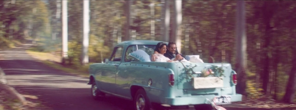 Newlyweds drive away in a vintage teal pickup truck, decorated with flowers, down a tree-lined rural road, post-wedding celebration.