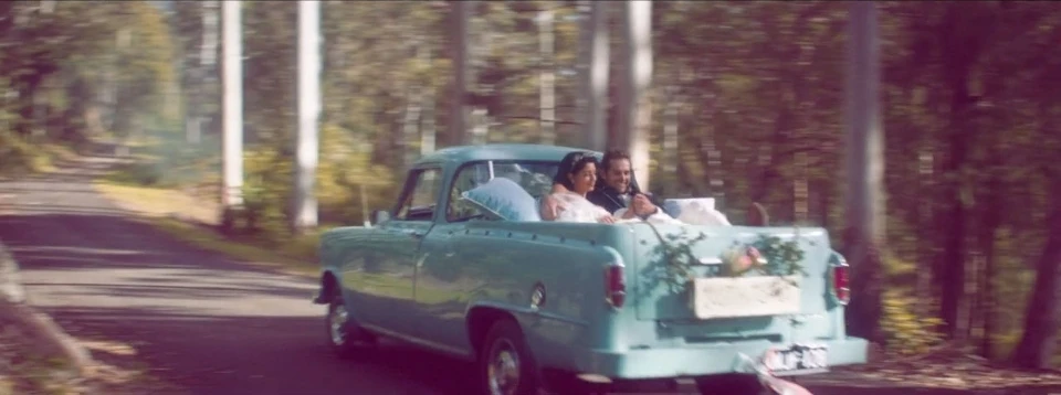 Newlyweds drive away in a vintage teal pickup truck, decorated with flowers, down a tree-lined rural road, post-wedding celebration.