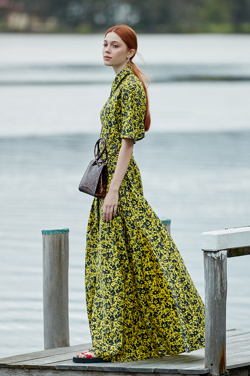 A red-haired woman in a yellow and black floral dress stands on a wooden dock with a brown purse, with a lake and trees in the background.