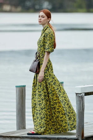 A red-haired woman in a yellow and black floral dress stands on a wooden dock with a brown purse, with a lake and trees in the background.