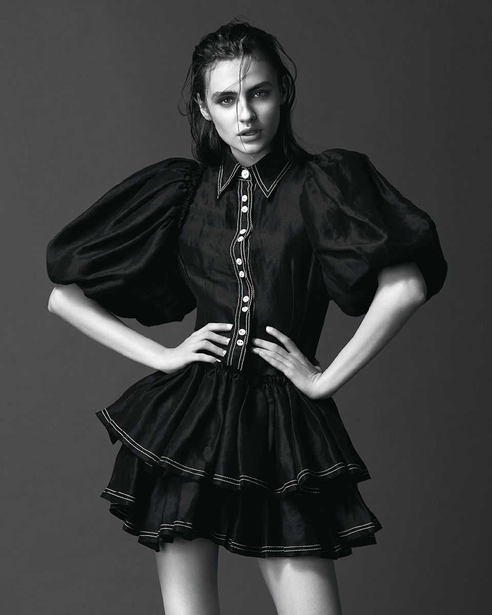 A young adult female model poses in a black dress with a tiered skirt and puffed sleeves, set against a neutral gray background.