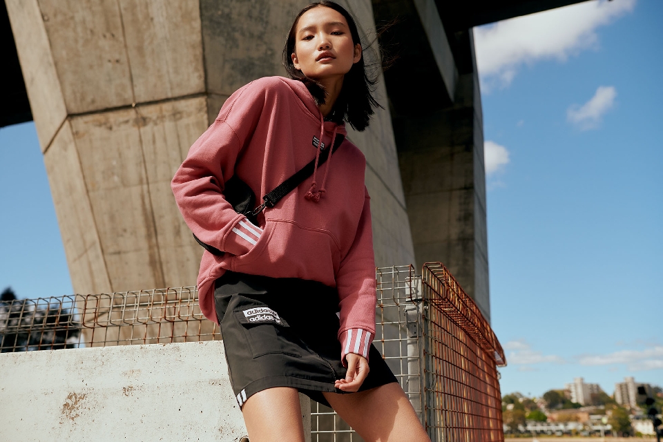 Asian woman in dusty rose hoodie and black skirt posing against concrete underpass with blue sky. Adidas apparel fashion shoot.
