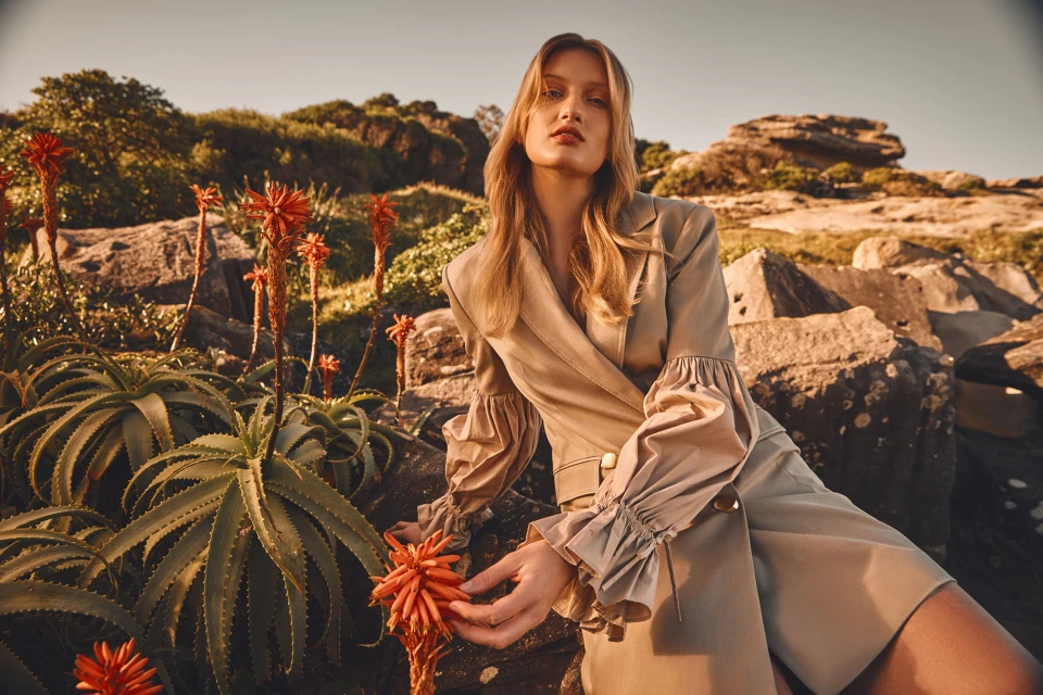 A woman in a beige dress poses among rocks and flowering plants. The image is brightly lit and has a warm, natural feel.