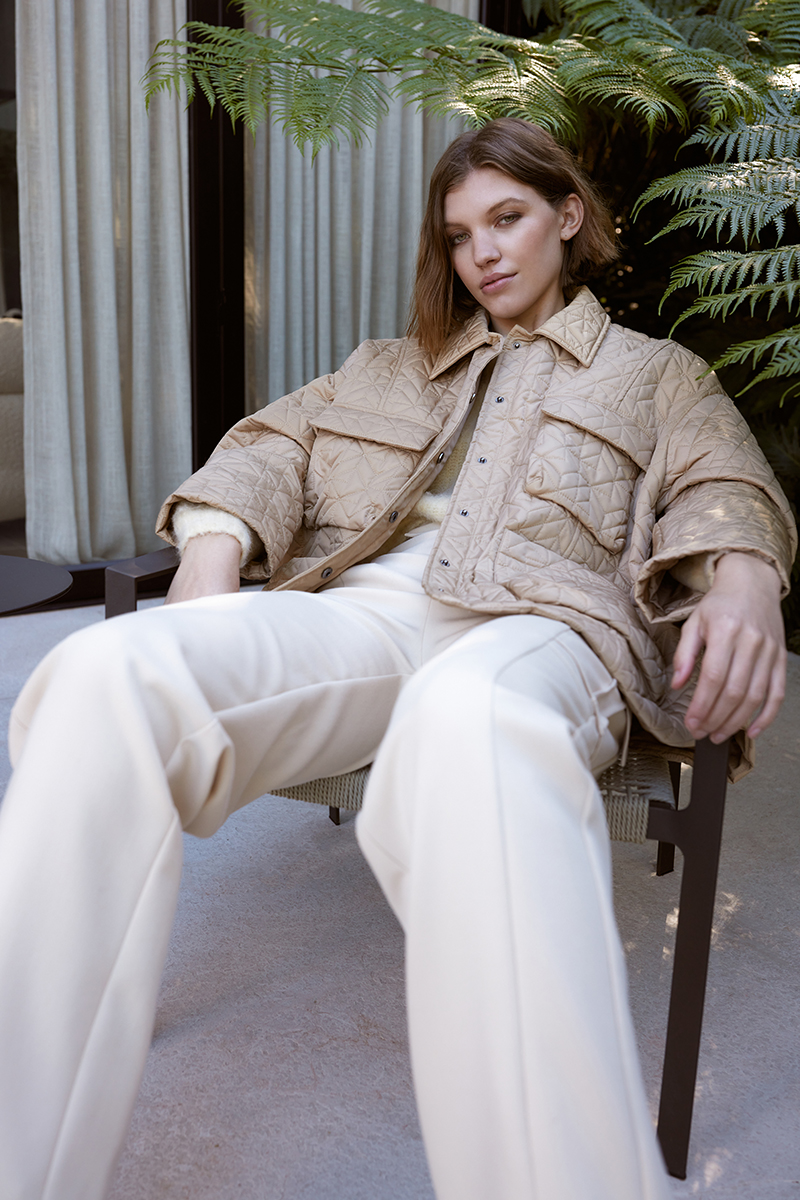 A woman sits in a chair wearing a quilted jacket and wide-leg pants in an outdoor setting with leafy green plants in the background.