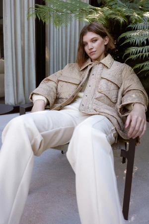 A woman sits in a chair wearing a quilted jacket and wide-leg pants in an outdoor setting with leafy green plants in the background.