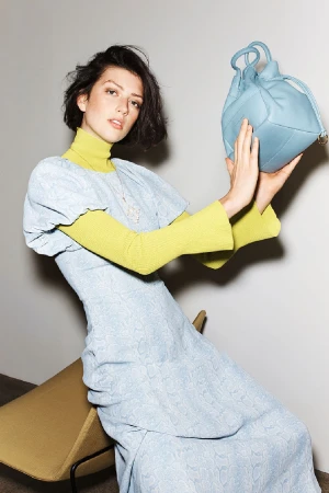 Model with dark short hair wearing a light blue dress over a yellow turtleneck, holding a blue leather handbag against a white wall.