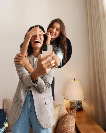 Two smiling women taking a selfie indoors, one wearing a houndstooth blazer and blue jeans while holding a smartphone