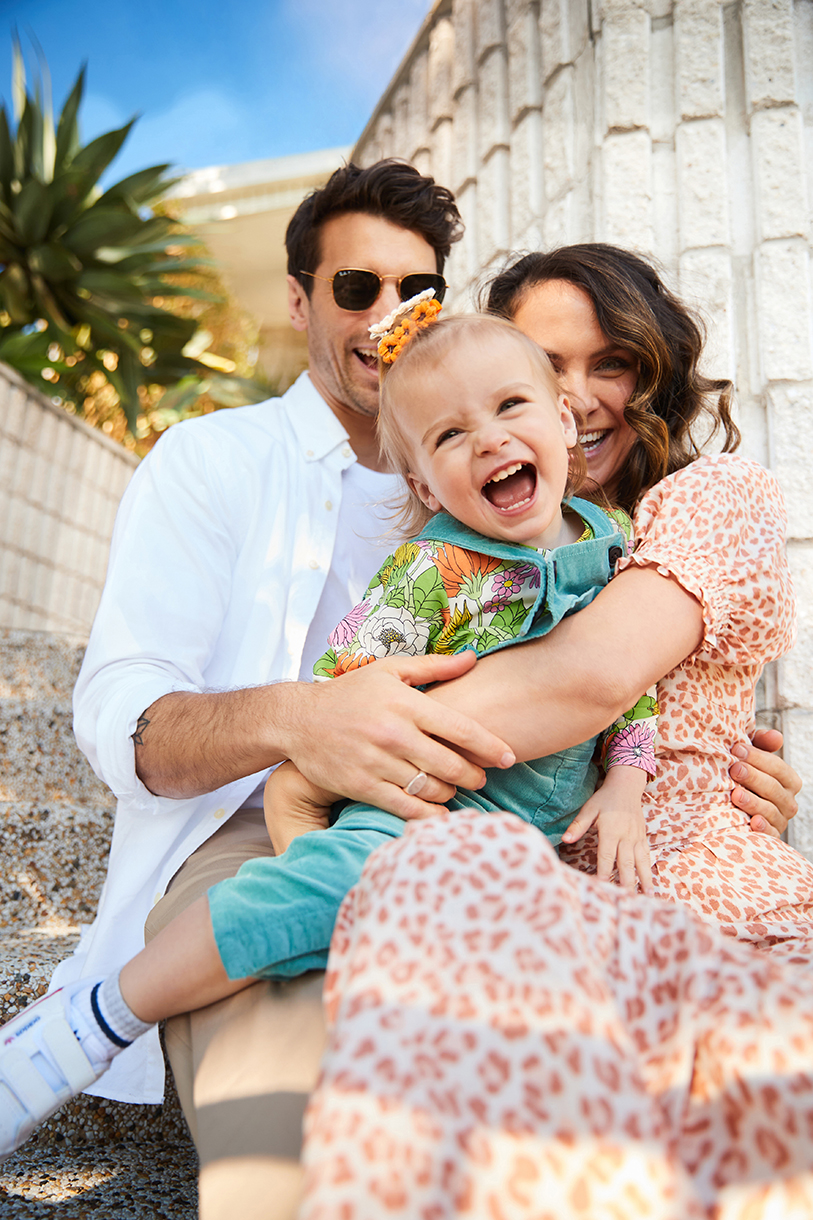 Family portrait on concrete steps featuring a smiling toddler in overalls held by her mother, with father standing close, bathed in sunlight.