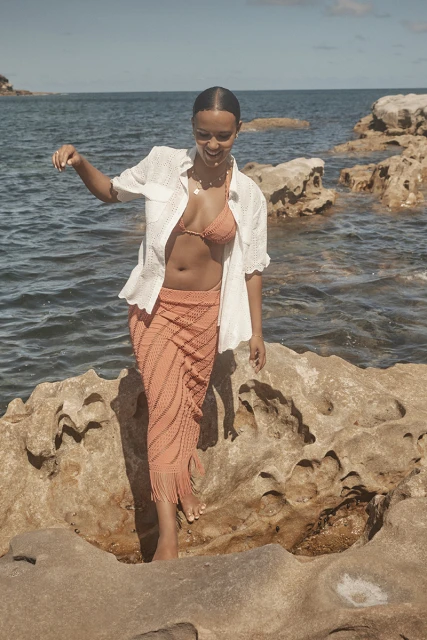 A young adult woman with tan skin walks on rocks by the ocean, wearing a knit skirt, bra top, and a white button-down shirt.