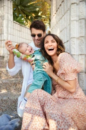 Family of three with a laughing toddler lifted in the air by a smiling man and a woman in a leopard-print dress
