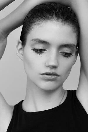 Close-up monochrome shot of a fair-skinned woman with slicked-back hair in a black top, posing with her arms raised above her head.