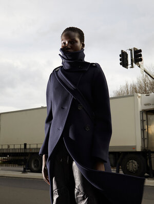 Fashion shot of a dark-skinned model wearing a blue coat with a high collar and a shoulder strap standing on a road in front of a truck.