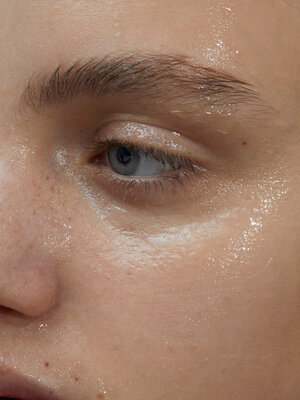Close-up of a young adult's face, focusing on a light blue eye, eyebrow, freckles, and glistening skin, capturing natural beauty.