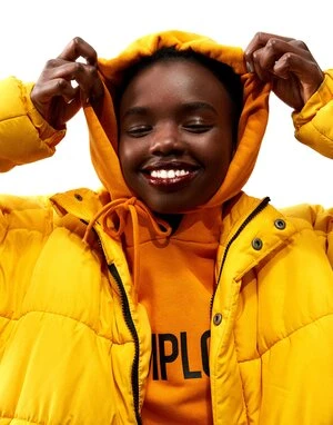 A dark-skinned woman smiles while wearing a yellow puffer jacket and a yellow hooded sweatshirt against a white backdrop in a studio setting.