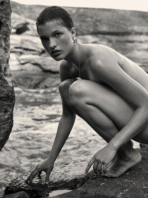 Black and white portrait of a young adult woman squatting near water, looking at the camera with rocks and water in the background.