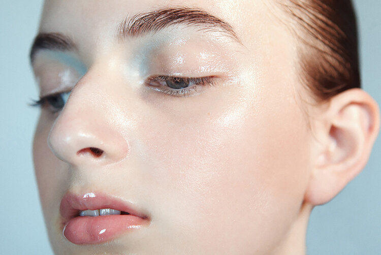 Close-up portrait of a young woman with glossy skin and lips. The shot focuses on her face, showcasing her fresh, dewy complexion.
