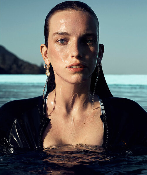 Adult female with wet hair and black wetsuit stands in water with mountains in the background. She wears gold earrings and natural makeup.