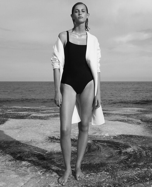 Woman in black swimsuit stands on rock formation at ocean's edge wearing a white jacket in black and white, fashion photography.