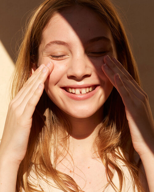 Smiling young woman with closed eyes touches her face in a close-up studio shot with warm lighting and a neutral background.