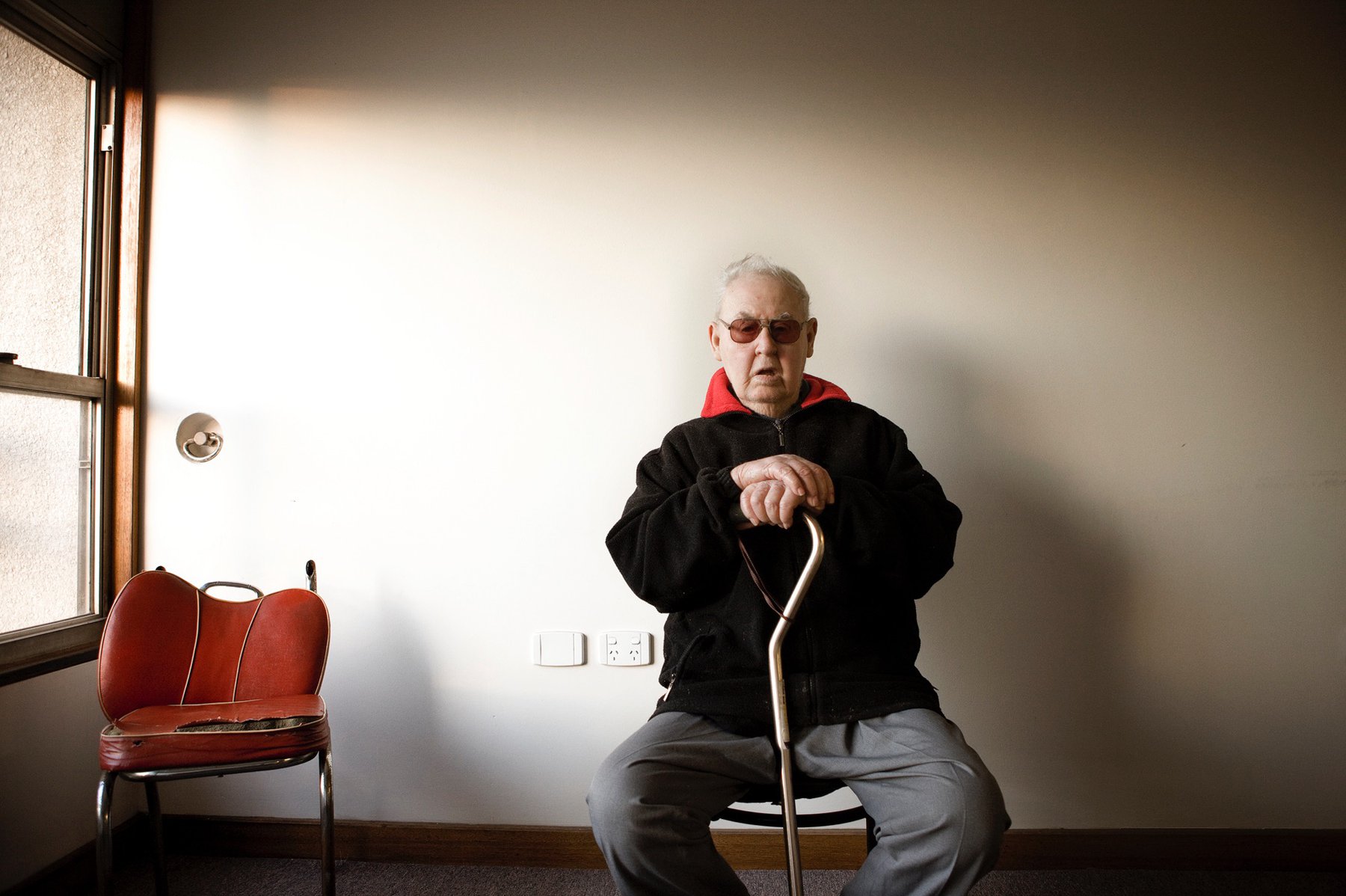 Elderly man with cane sits in a room near a window next to an old chair, wearing glasses and a dark jacket over a red shirt.