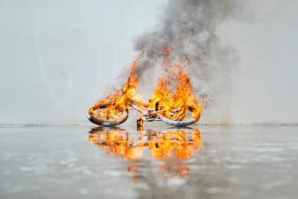 A bicycle engulfed in flames with smoke against a light gray backdrop reflecting on a wet surface, creating a dramatic and surreal image.