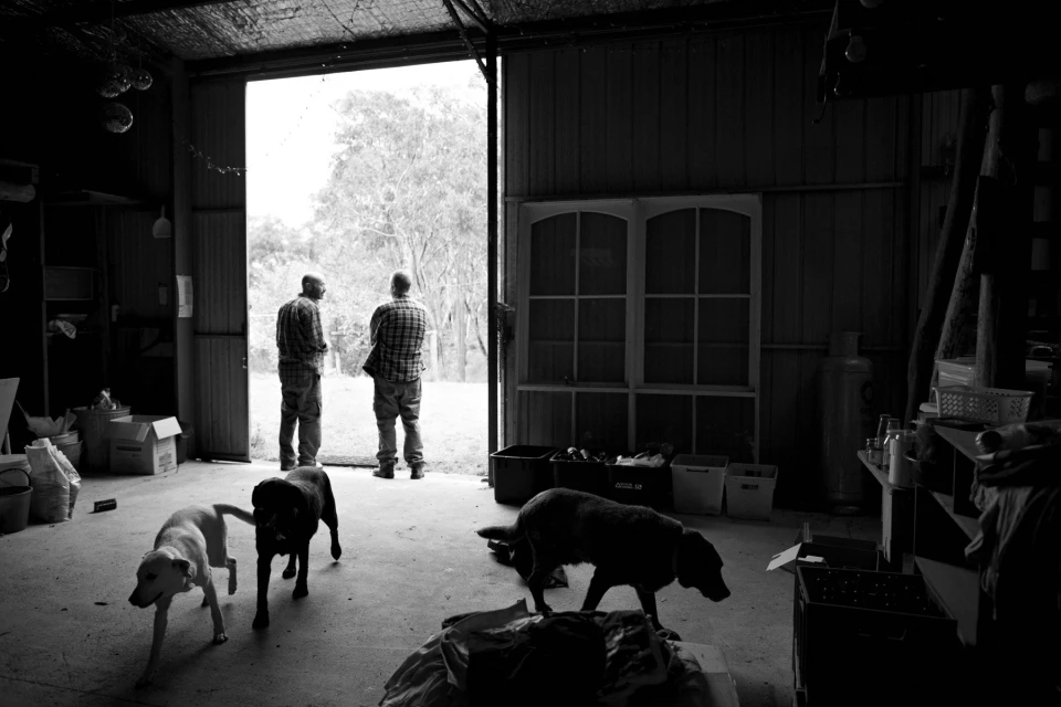 Two men standing at the entrance of a garage, with three dogs in the foreground, in a monochromatic, indoor setting with natural light.