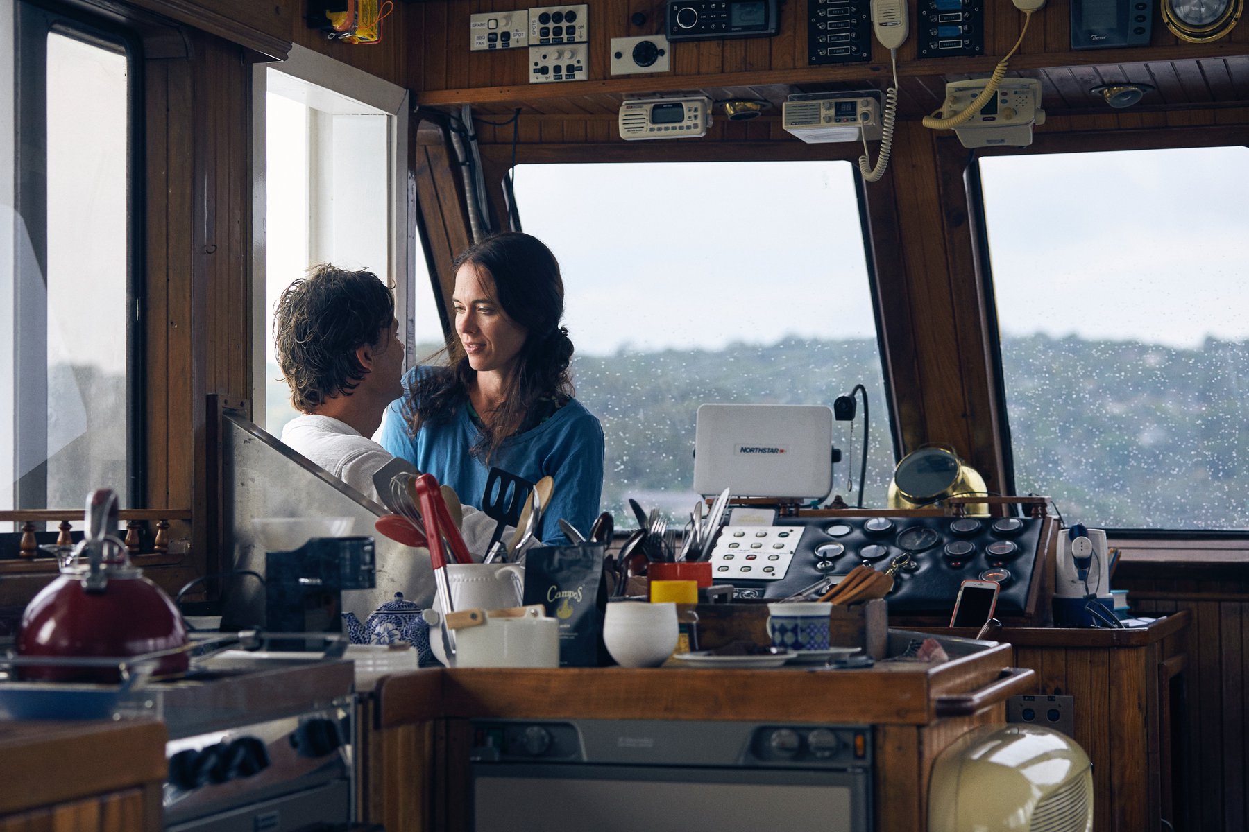 A serene scene inside a boat, featuring a couple near the helm, surrounded by cooking utensils and nautical equipment against a window backdrop.