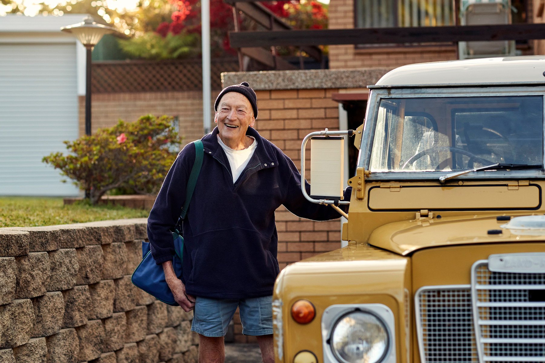 Elderly man smiles standing next to an old yellow car in front of a brick house, wearing a dark jacket, hat, and carrying a blue bag.