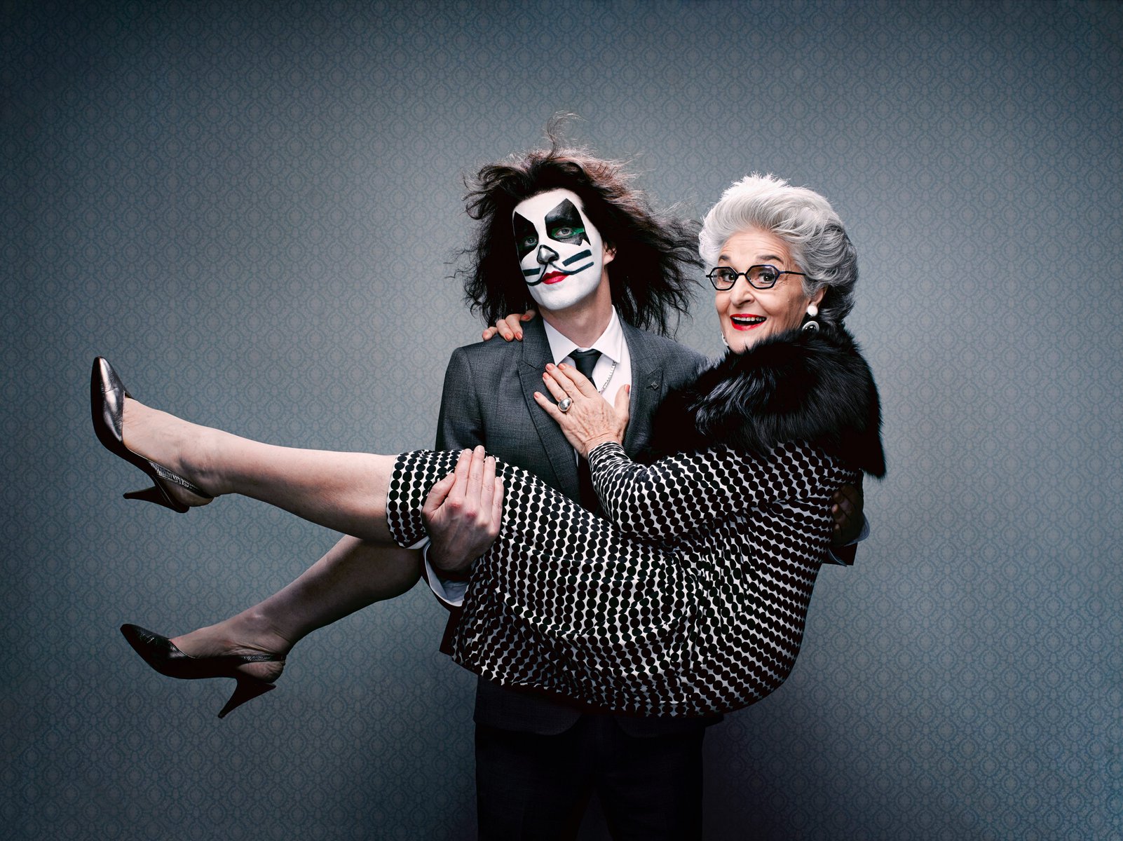 Young man in face paint carrying an elderly woman in a polka dot dress, in a studio setting with a patterned background. Happy expression.