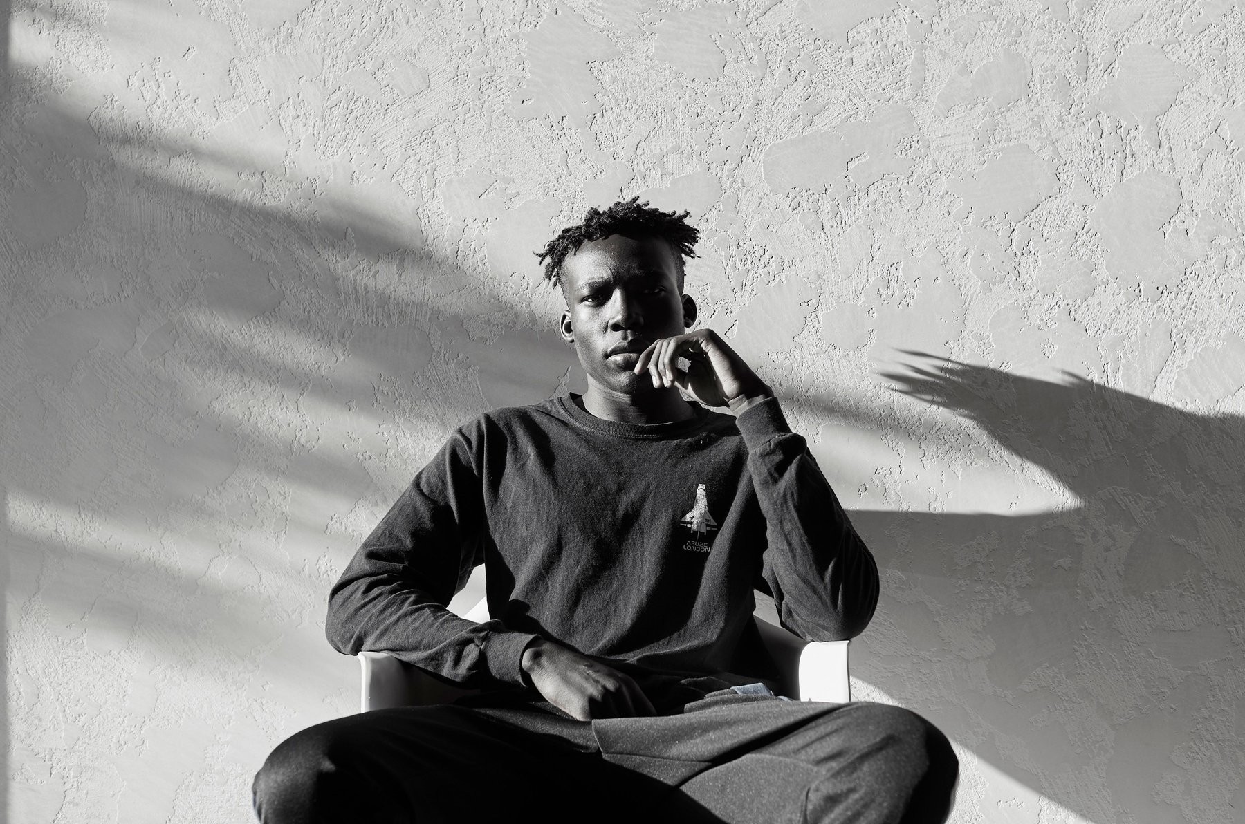 Monochrome portrait of a young African American man in long-sleeve shirt sitting on a white chair indoors with light and shadow play.