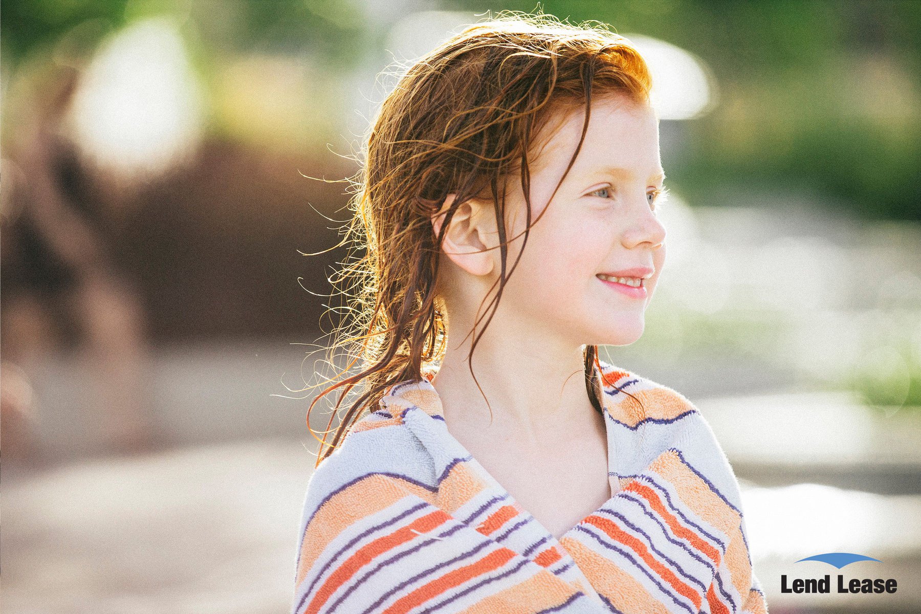 Portrait of a smiling young girl with wet red hair wrapped in a striped towel outdoors in soft sunlight, with blurred background.