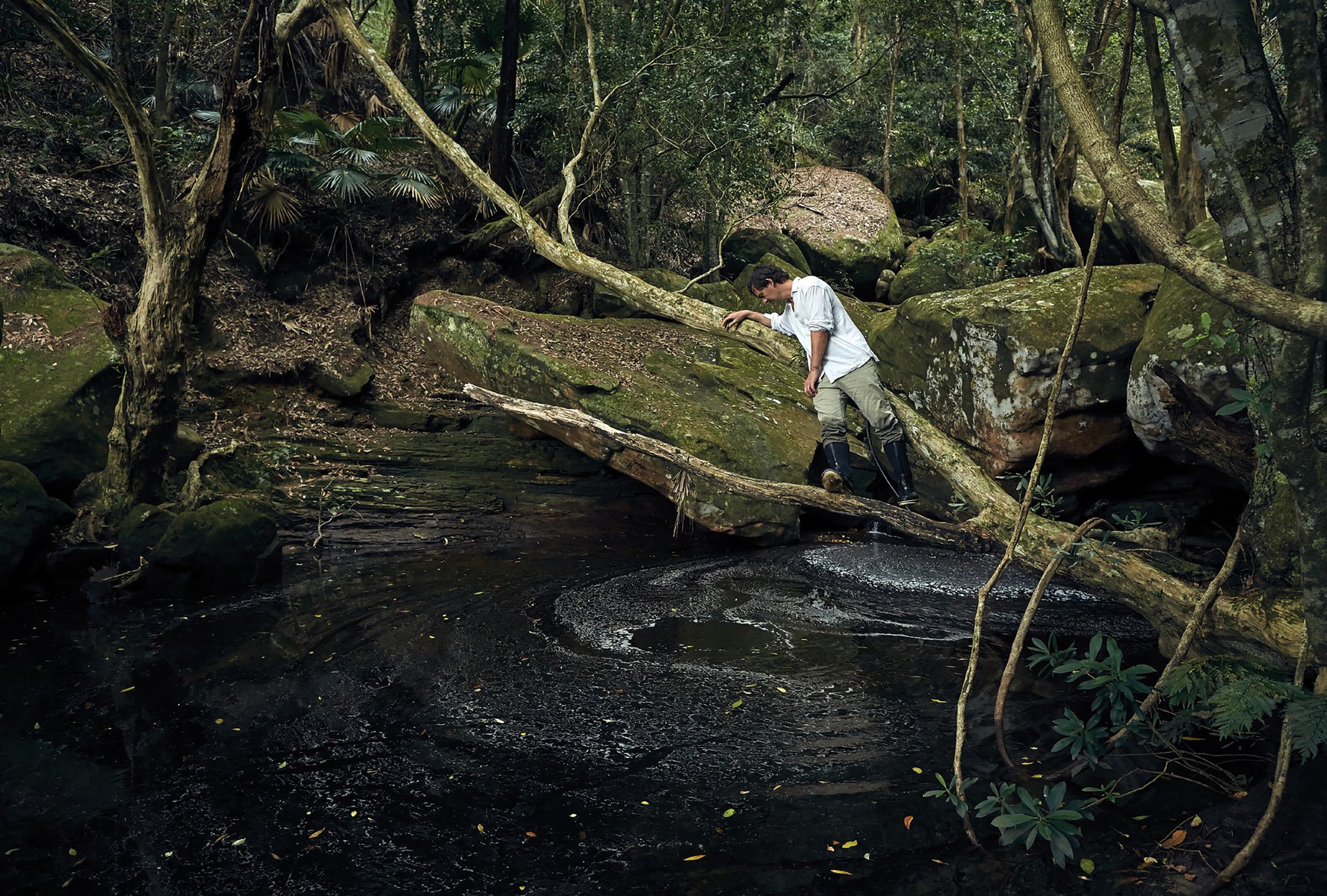 Man in white shirt and khaki pants carefully crosses a dark creek on a fallen log in a lush green forest with mossy rocks.