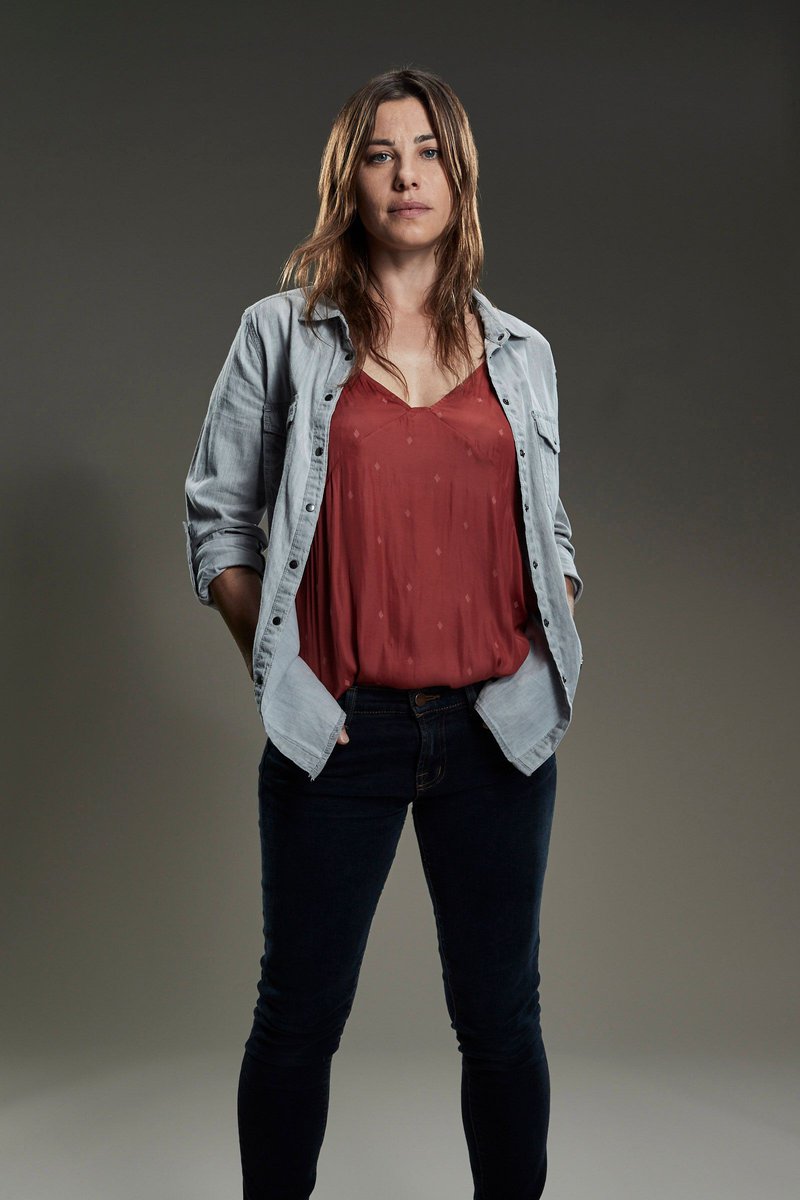 Full-length portrait of a woman with long brown hair wearing a red tank top, light blue shirt, and dark blue jeans, standing indoors.