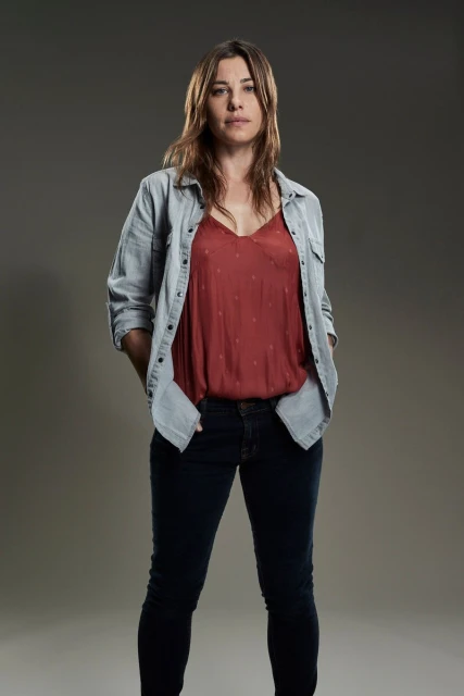 Full-length portrait of a woman with long brown hair wearing a red tank top, light blue shirt, and dark blue jeans, standing indoors.