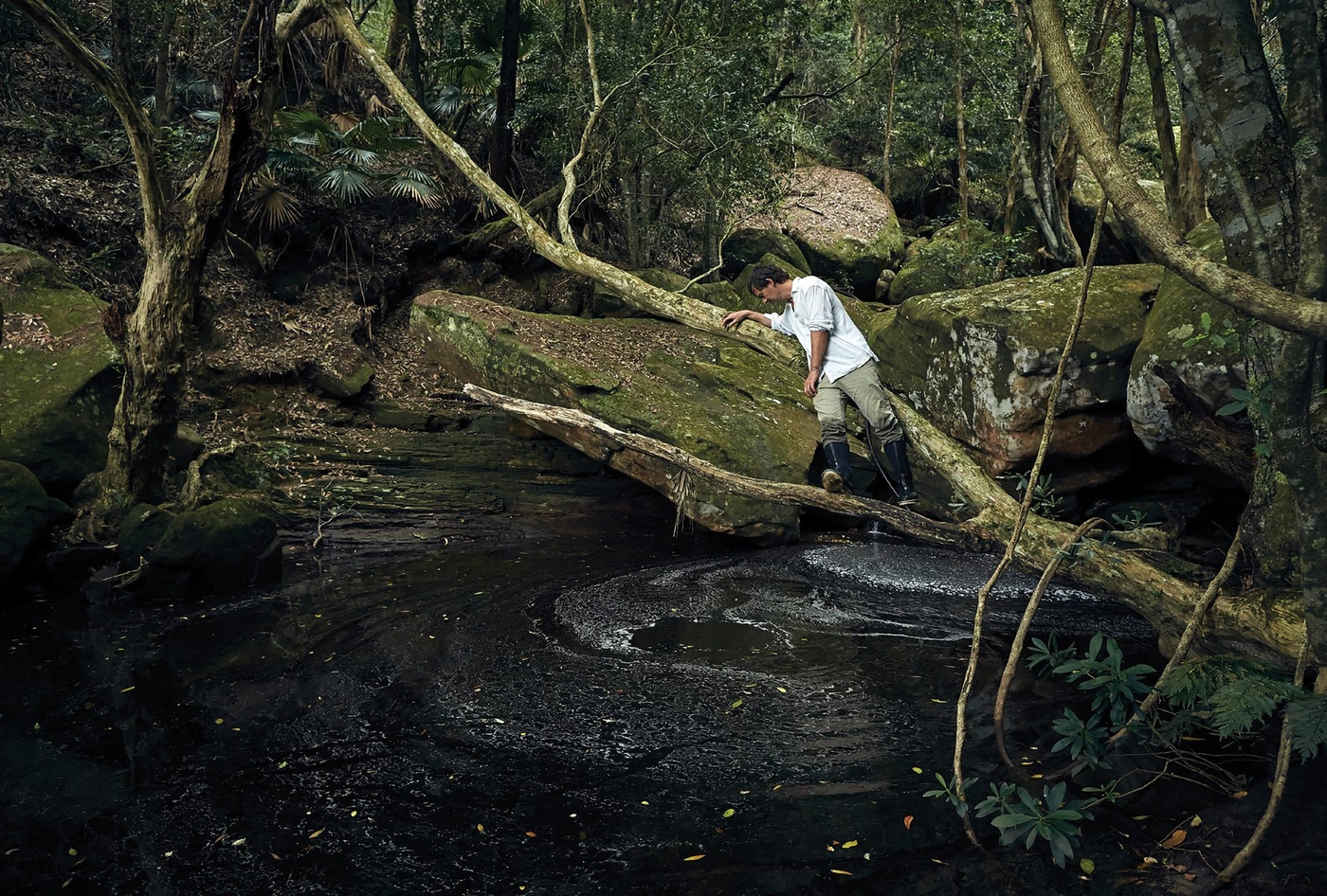 A man in a white shirt and trousers leans against a fallen branch over a dark forest pool, photographed by Toby Burrows.