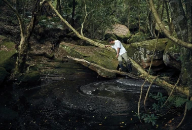 A man in a white shirt and trousers leans against a fallen branch over a dark forest pool, photographed by Toby Burrows.